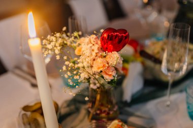 Bir mum, kalp şeklinde bir süs, pembe çiçekler ve cam bir vazoda bebek nefesiyle süslenmiş bir masa.