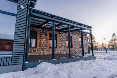 Günbatımında ağaçlı ve berrak gökyüzü olan karlı bir arazide kurulu, ahşap bir veranda ve geniş pencereler modern taş yapıya sahiptir.