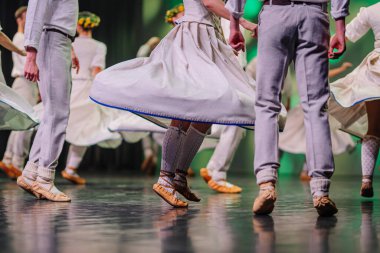 Geleneksel giyinmiş Letonyalı halk dansçıları, kültürel bir etkinlik sırasında girdaplı etekleri ve detaylı ayakkabıları vurgulayarak sahnede gösteri yapıyorlar.