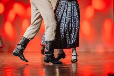 Letonyalı halk dansçıları sahnede dans ederken geleneksel kıyafetlerini sergiliyorlar, parlak ahşap bir zemin ve canlı kırmızı bir zemin