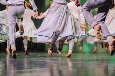 Geleneksel giysiler içindeki Letonyalı halk dansçıları, renkli kostümler ve dinamik ayak hareketlerini vurgulayarak sahnede enerjik dans adımları atıyorlar.
