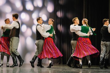 Valmiera, Letonya - 27 Aralık 2025 Letonya Halk Dansı Konseri. Canlı kostümler içindeki Letonyalı halk dansçıları, yumuşak ışıklandırılmış arka planlarıyla sahnede geleneksel dans figürlerini sergiliyorlar.