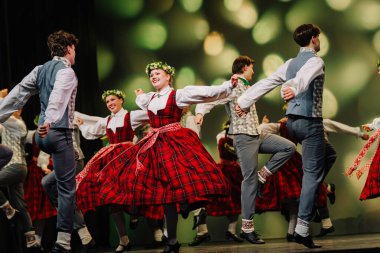 Valmiera, Letonya - 27 Aralık 2025 Letonya Halk Dansı Konseri. Letonyalı halk dansçıları arka planda renkli ışıklandırmalarla sahneye çıkarken geleneksel kıyafetlerini sergiliyorlar
