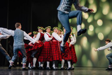 Valmiera, Letonya - 27 Aralık 2025 Letonya Halk Dansı Konseri. Renkli kostümlü Letonyalı halk dansçıları parlak ışıklar ve nişanlı bir seyirciyle yeteneklerini sahnede sergiliyorlar.