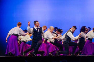 Valmiera, Letonya - 27 Aralık 2025 Letonya Halk Dansı Konseri. Letonyalı halk dansçıları, sahne performansları sırasında mor etekli ve beyaz gömlekli geleneksel giysiler sergilediler.