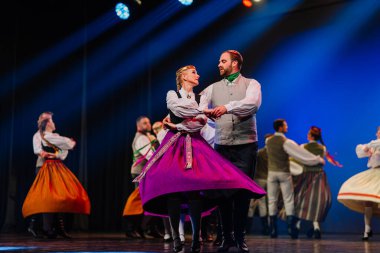Valmiera, Letonya - 27 Aralık 2025 Letonya Halk Dansı Konseri. Geleneksel giyinmiş Letonyalı halk dansçıları renkli ışıklandırmalarla sahnede canlı bir performans sergilerler.
