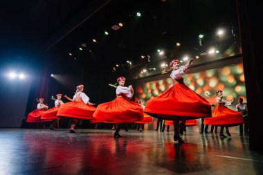 Valmiera, Letonya - 27 Aralık 2025 Letonya Halk Dansı Konseri. Letonyalı halk dansçıları sahnede parlak ışıklar ve geleneksel kıyafetlerle kırmızı etekler içinde dönerler.