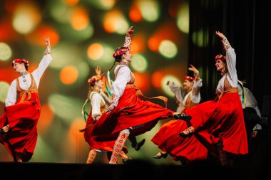 Valmiera, Letonya - 27 Aralık 2025 Letonya Halk Dansı Konseri. Kırmızı ve beyaz kostümlü Letonyalı halk dansçıları dans hareketlerini renkli ışıklandırmalarla sahnedeler.