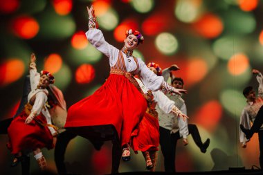 Valmiera, Letonya - 27 Aralık 2025 Letonya Halk Dansı Konseri. Geleneksel giyinmiş Letonyalı halk dansçıları sahneyi aydınlatan renkli ışıklarla sahnede gösteri yapıyorlar.
