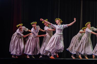 Valmiera, Letonya - 27 Aralık 2025 Letonya Halk Dansı Konseri. Beyaz elbiseli ve çiçekli taç giyen Letonyalı halk dansçıları sahnede zarif bir şekilde hareket ediyorlar.