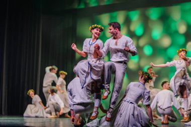 Valmiera, Letonya - 27 Aralık 2025 Letonya Halk Dansı Konseri. Geleneksel giyinmiş Letonyalı halk dansçıları canlı ışıklandırma ve çiçeklerle sahnede gösteri yapıyorlar.