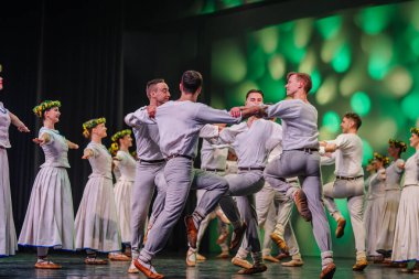 Valmiera, Letonya - 27 Aralık 2025 Letonya Halk Dansı Konseri. Geleneksel giyinmiş Letonyalı halk dansçıları canlı ışıklandırma ve çiçeklerle sahnede gösteri yapıyorlar.
