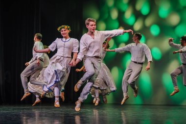 Valmiera, Letonya - 27 Aralık 2025 Letonya Halk Dansı Konseri. Geleneksel giyinmiş Letonyalı halk dansçıları arkalarında renkli ışıklandırmalarla zarif bir şekilde sahneye atlıyorlar.