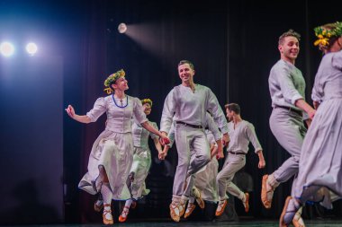 Valmiera, Letonya - 27 Aralık 2025 Letonya Halk Dansı Konseri. Geleneksel giyinmiş Letonyalı halk dansçıları sahneyi aydınlatan parlak ışıklarla neşe içinde sahnede gösteri yapıyorlar.