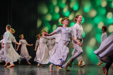 Valmiera, Letonya - 27 Aralık 2025 Letonya Halk Dansı Konseri. Geleneksel giyinmiş Letonyalı halk dansçıları arkalarında renkli ışıklandırmalarla sahnede zarifçe ilerliyorlar.