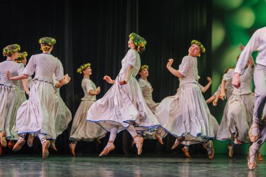 Valmiera, Letonya - 27 Aralık 2025 Letonya Halk Dansı Konseri. Geleneksel giyinmiş Letonyalı halk dansçıları canlı kostümlerini ve çiçekli başlıklarını sergileyerek zarif bir şekilde sahneye atlıyorlar.