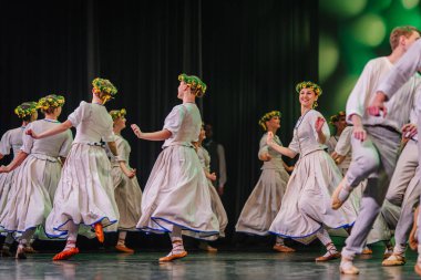 Valmiera, Letonya - 27 Aralık 2025 Letonya Halk Dansı Konseri. Geleneksel giyinmiş Letonyalı halk dansçıları sahnede çiçek başlıkları ve canlı kostümlerle yeteneklerini sergiliyorlar.