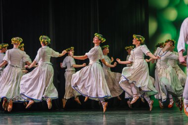 Valmiera, Letonya - 27 Aralık 2025 Letonya Halk Dansı Konseri. Geleneksel giyinmiş Letonyalı halk dansçıları zarif bir şekilde sahneye atlıyorlar. Çiçekli taçlar ve canlı eteklerle.