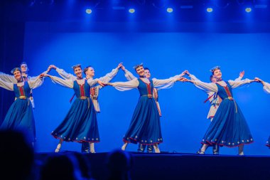 Valmiera, Letonya - 27 Aralık 2025 Letonya Halk Dansı Konseri. Mavi geleneksel kostümlü dansçılar mavi arka plan ışıklandırmasıyla sahnede şarkı söylerken el ele tutuşuyorlar.