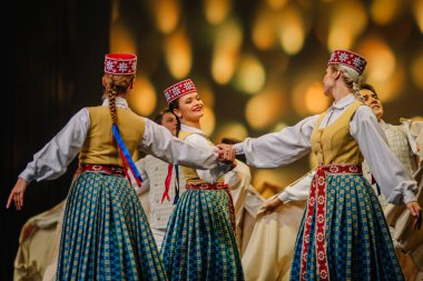 Valmiera, Letonya - 27 Aralık 2025 Letonya Halk Dansı Konseri. Geleneksel Letonya kıyafetli üç kadın sahnede dans ederken arkalarında parlak ışıklar bırakarak el ele tutuşuyorlar.