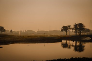 Dubai, Birleşik Arap Emirlikleri - 13 Ocak 2026: Güneşin doğuşunda golf sahasında palmiye ağaçları ve arka planda bir şehir silüeti bulunan yansıtıcı bir su kütlesi