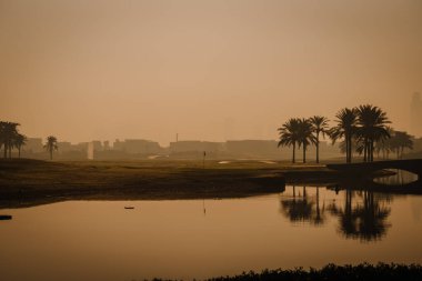 Dubai, Birleşik Arap Emirlikleri - 13 Ocak 2026: Güneşin doğuşunda golf sahasında palmiye ağaçları ve arka planda bir şehir silüeti bulunan yansıtıcı bir su kütlesi