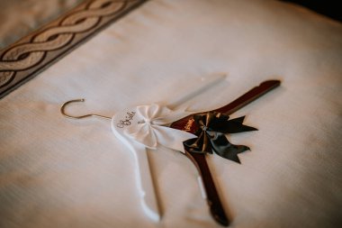 Gelin askısı beyaz kurdele ve siyah fiyonkla süslenmiş, üzerinde düğün temalı betimlemeler için uygun desenli kumaş bir yüzeye yerleştirilmiş 