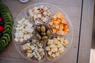 Çedar, gouda ve yeşil zeytinli mavi peyniri de içeren çeşitli peynir küpleri ahşap bir yüzey üzerinde şeffaf bir tepside dizilmiş.