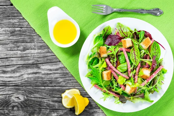 Délicieuse salade fraîche de salami et feuilles de laitue mélangées - bébé épinards, roquette, blette dans un plat blanc sur la vieille table en bois, vue sur le dessus — Photo