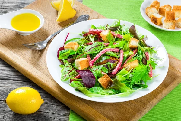 Deliciosa salada fresca de salame e folhas de alface mistas - espinafre de bebê, rúcula, acelga em um prato branco na velha mesa de madeira, vista superior — Fotografia de Stock