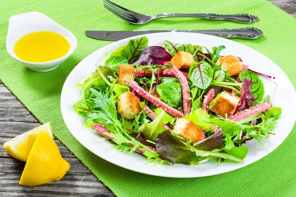 Deliciosa salada fresca de salame e folhas de alface mistas - espinafre de bebê, rúcula, acelga em um prato branco na velha mesa de madeira, vista superior — Fotografia de Stock