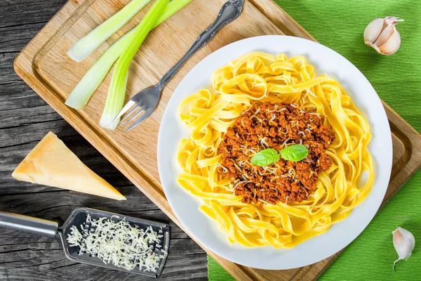 Bolognese ragout with italian pasta on a white plate, decorated with basil leaves, authentic recipe, wooden background with celery, garlic, cherry tomatoes and parmesan cheese, full focus, close-up — Stock Photo, Image