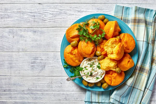 Southern cuisine: gluten-free corn muffins of cornmeal, yogurt, and eggs served on a blue plate with cream cheese, fresh parsley, and green olives on a white wooden background, top view, close-up