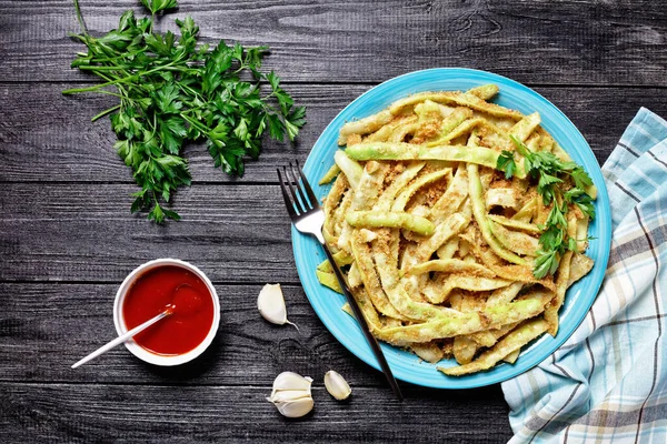 Golden french low-calorie wax beans with garlic and parsley coated with breadcrumbs and fried on a skillet, served on a blue plate with fork on a dark wooden table, top view, close-up