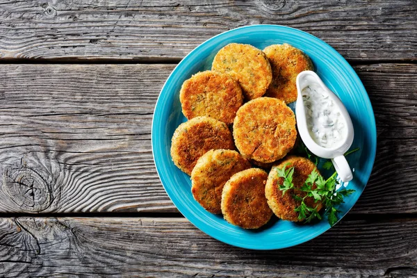 Easy fish cakes of white fish fillet: cod or haddock with potato and parsley, breaded in breadcrumbs served on a plate with tartar sauce in a gravy boat on a wooden background,  top view, close-up
