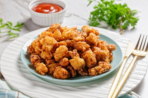 Popcorn chicken - american fast food dish of bite-sized chicken pieces breaded and fried, served on a white plate with ketchup and golden fork on a white marble stone background, top view, close-up