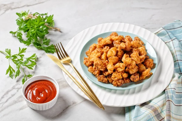 Popcorn chicken - american fast food dish of bite-sized chicken pieces breaded and fried, served on a white plate with ketchup and golden fork on a white marble stone background, top view, close-up