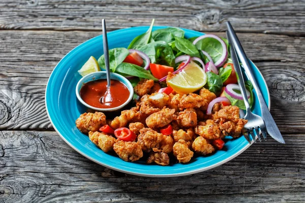 Crispy popcorn chicken american snack served on a blue plate with ketchup and salad of baby spinach, red onion, cherry tomatoes and lemon wedges on a wooden table, top view, close-up