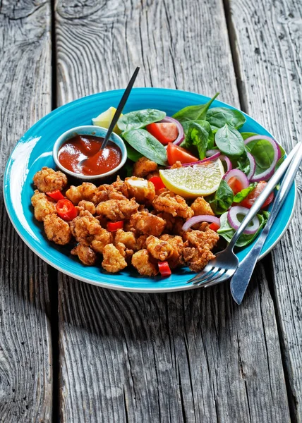 Crispy popcorn chicken american snack served on a blue plate with ketchup and salad of baby spinach, red onion, cherry tomatoes and lemon wedges on a wooden table, top view, close-up