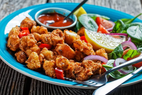 Crispy popcorn chicken american snack served on a blue plate with ketchup and salad of baby spinach, red onion, cherry tomatoes and lemon wedges on a wooden table, top view, close-up