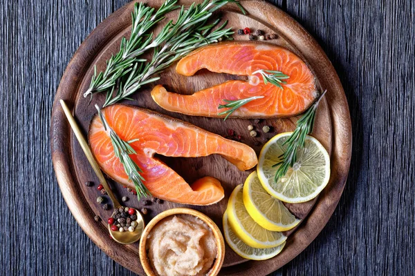 Smoked arctic char steaks on a round cutting board on a dark wooden background with cod roe, lemon, fresh rosemary, peppercorn, top view, close-up — Stock Image Smoked Arctic Char Steaks Cutting Board Dark Wooden Background Cod Stock Picture