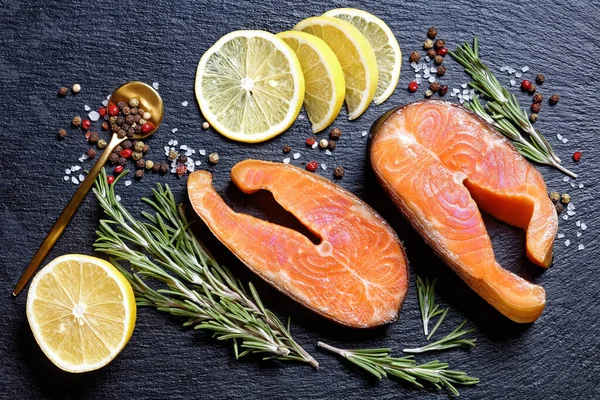Smoked arctic char steaks on a round cutting board on a dark slate background with lemon, fresh rosemary, peppercorns, top view, close-up — Stock Image Smoked Arctic Char Steaks Cutting Board Dark Slate Background Lemon Stock Image