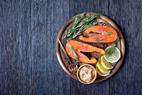 Smoked arctic char steaks on a round cutting board on a dark wooden background with cod caviar, lemon, fresh rosemary, peppercorn, top view, close-up, copy space — Stock Image Smoked Arctic Char Steaks Cutting Board Dark Wooden Background Cod Royalty Free Stock Images