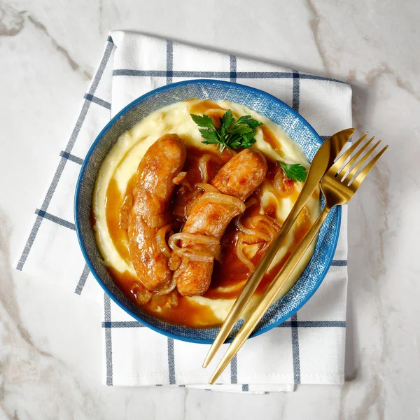 British food bangers and mash or pork sausage with onion gravy and mashed potato on a plate with golden cutlery on a marble table, top view, close-up