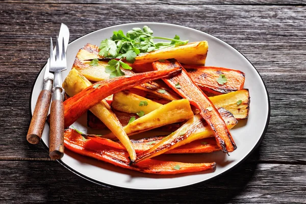 Moroccan roasted root vegetables: parsnip and carrot with ras el hanout spice: cinnamon, turmeric, anise served on a plate with fresh coriander and cutlery on a wooden background, top view, close-up