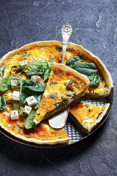 feta spinach cheddar cheese quiche in a baking dish on a wooden board on a grey concrete table with a slice on a spatula, vertical view from above,  french cuisine