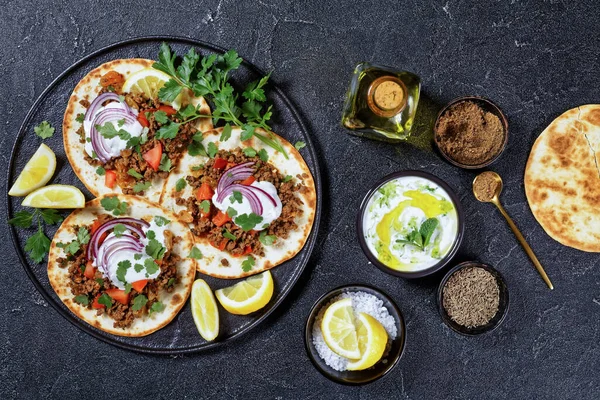 ground Lamb Flatbreads topped with yogurt, coriander leaves and red onion on a black platter on a concrete table, top view, flat lay