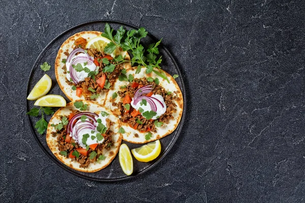 ground Lamb Flatbreads topped with yogurt, coriander leaves and red onion on a black platter on a concrete table, top view, flat lay, free space