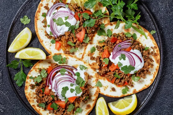 ground Lamb Flatbreads topped with yogurt, coriander leaves and red onion on a black platter on a concrete table, flat lay, macro