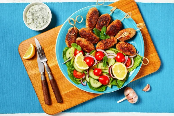 Lamb kofta kebabs of ground lamb, served on a blue plate with yogurt dressing, fresh vegetables salad, and lemon wedges, greek cuisine on a white wooden table, top view, close-up